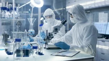 Güvenli yüksek düzey laboratuvar bilim adamları bir Coverall içinde yapılan bir araştırma. Kimyager bir petri kerpeten ile örnekleri ayarlar ve sonra onları mikroskop altında inceler ve meslektaşı sonuçları yazıyor.