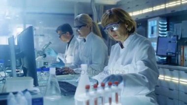 Kadın ve erkek bilim adamları çalışma onların bilgisayar içinde büyük Modern Laboratuvar hakkında. Kadehler, kimyasallar ve farklı teknik ekipmanlar ile çeşitli raflarına nedir görünür.