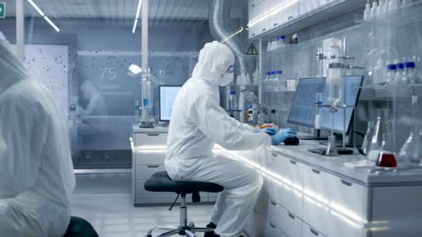 In a Secure High Level Laboratory Scientists in a Coverall Conducting a Research. Biólogo ajusta muestras en un plato de Petri con pinzas y luego las examina bajo el microscopio y su colega analiza los resultados en una computadora . — Vídeos de Stock