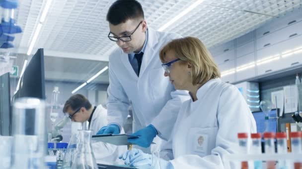 In Modern Laboratory Senior Female Scientist Has Discussion with Young Male Laboratory Assistant. Muestra sus gráficos de datos en un portapapeles, lo analiza y lo ingresa en su computadora . — Vídeo de stock