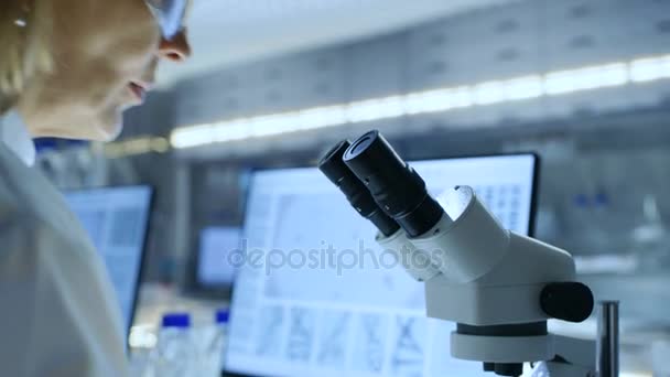 Bióloga Sênior trabalha em seu computador e olha para os materiais sob microscópio. Ela está num laboratório moderno. . — Vídeo de Stock