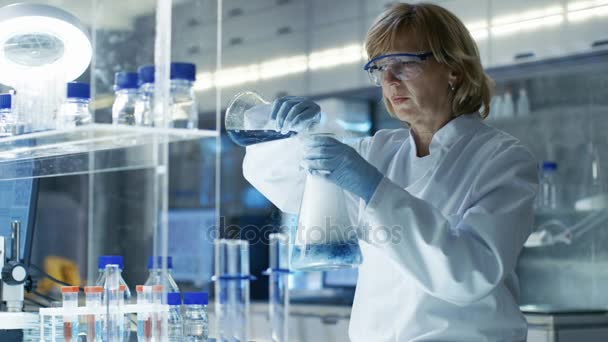 Senior Female Chemist in Safety Glasses Mixes Smoking Liquids in a Beakers. Elle travaille dans un laboratoire moderne lumineux avec des étagères pleines d'échantillons, de tubes à essai et d'équipements technologiques de pointe . — Video