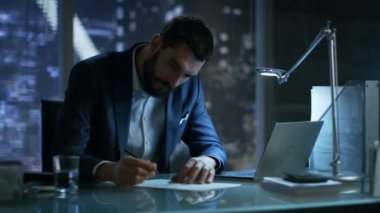 Gece geç saatlerde işadamı büyük şehir pencere manzaralı özel ofisinde bir dizüstü bilgisayar ve işaretler belgeler üzerinde çalışır.