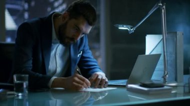 Gece geç saatlerde işadamı büyük şehir pencere manzaralı özel ofisinde bir dizüstü bilgisayar ve işaretler belgeler üzerinde çalışır.