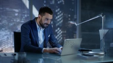 Özel Office işadamı kamera üzerindeki bir Laptop gülümsüyor üzerinde çalışıyor, gece geç saatlerde. Arka plan büyük şehir pencere görünümünde.