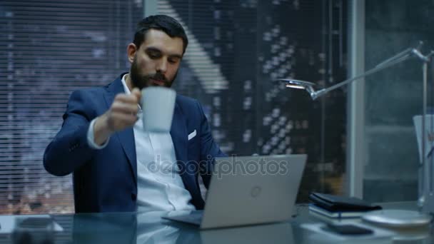 Tarde en la noche Empresario sobrecargado de trabajo trabaja en un ordenador portátil y bebe café en su oficina privada con vista a la ventana de la gran ciudad . — Vídeos de Stock