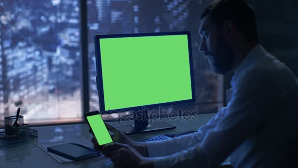 Tarde en la noche empresario trabaja en un ordenador y Smartphone con pantalla verde maqueta en su oficina privada con gran ciudad de la ventana. — Vídeos de Stock