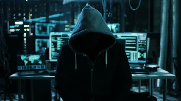Dangerous Internationally Wanted Hacker with Covered Face Habla en la cámara. En segundo plano su sala de operaciones con múltiples pantallas y cables . — Vídeos de Stock