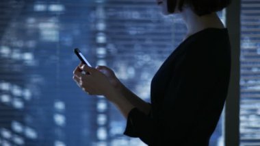 Çekici esmer bir sıkı elbise kullanır Smartphone pencere yanında duran büyük şehir manzaralı süre içinde. Onun yerine karanlık görünüyor.