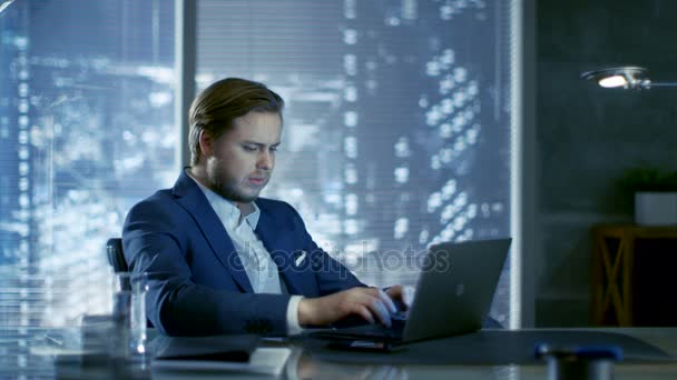 Succesvolle jonge zakenman zit in een kabinet op zijn bureau en werken op een Laptop. Achter hem venster met uitzicht op de grote stad. — Stockvideo