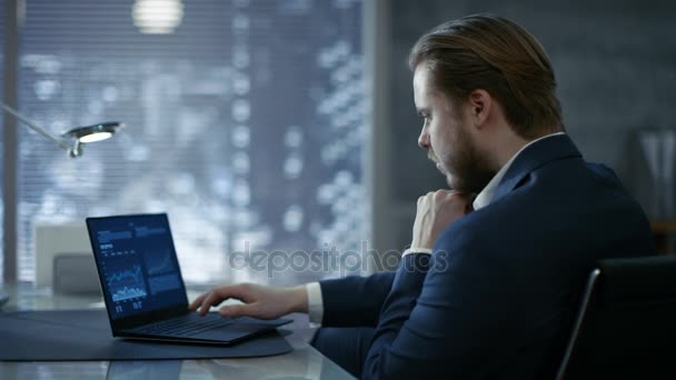 Jovem empresário bem sucedido senta-se em um escritório privado em sua mesa e trabalha em um laptop. Seu escritório é feito em estilo minimalista, é bastante escuro e tem janela com vista para a cidade grande . — Vídeo de Stock