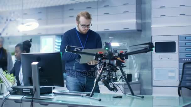 Jovem Engenheiro Programa Drone enquanto segura Laptop em suas mãos. Ele trabalha em um laboratório de alta tecnologia moderno brilhante . — Vídeo de Stock