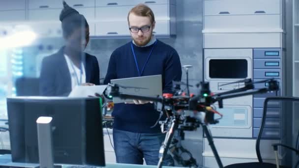 Ingenieros caucásicos masculinos y femeninos negros que trabajan en un proyecto de drones con ayuda de computadoras portátiles y tomando notas. Trabaja en un brillante laboratorio moderno de alta tecnología . — Vídeo de stock