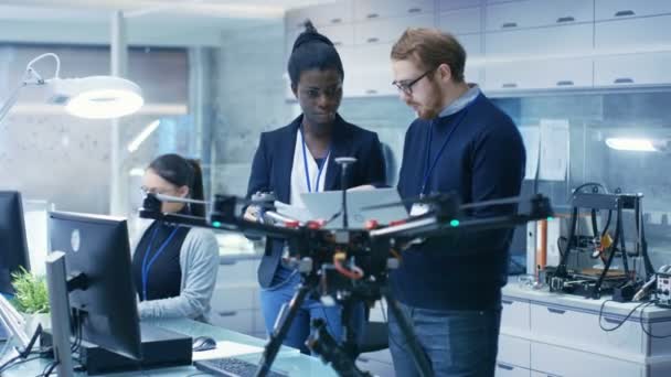 Ingenieros caucásicos masculinos y femeninos negros que trabajan en un proyecto de drones con ayuda de computadoras portátiles y tomando notas. Trabaja en un brillante laboratorio moderno de alta tecnología . — Vídeo de stock