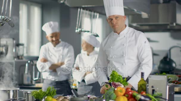 著名的一家大餐厅的厨师准备菜肴和相机上的微笑。在背景两个学徒和现代厨房. — 图库视频影像
