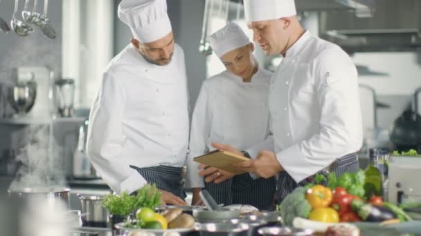 En el equipo de cocina moderna de los cocineros utilizan Tablet Computer para recetas, sonríen y tienen discusión. La cocina está llena de ingredientes alimentarios, verduras, carne, sopa hirviendo . — Vídeo de stock