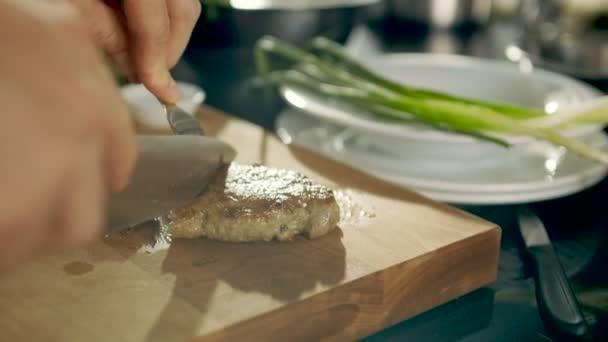 Primer plano de la pieza de carne preparada que se corta con el uso de cuchillo y tenedor en una tabla de cortar. En segundo plano Verduras verdes son visibles . — Vídeos de Stock