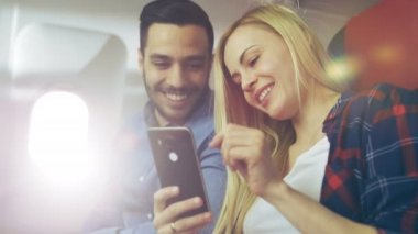Ticari uçak bir gemide güzel genç sarışın yakışıklı İspanyol erkek ile Smartphone ve gülümseme videoları izle. Uçak pencereden güneş parlıyor.