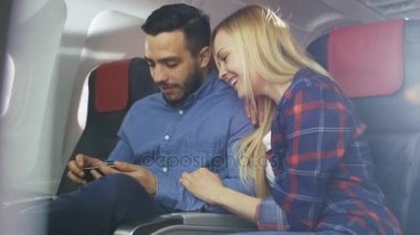 Ticari uçak bir gemide güzel genç sarışın yakışıklı İspanyol erkek ile Smartphone ve gülümseme videoları izle. Uçak pencereden güneş parlıyor.