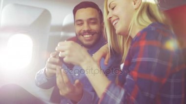 Ticari uçak bir gemide güzel genç sarışın yakışıklı İspanyol erkek ile Smartphone ve gülümseme videoları izle. Uçak pencereden güneş parlıyor.