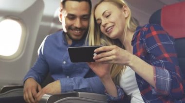 Ticari uçak bir gemide güzel genç sarışın yakışıklı İspanyol erkek ile Smartphone ve gülümseme videoları paylaşın. Uçak pencereden güneş parlıyor.