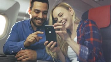 Ticari uçak bir gemide güzel genç sarışın yakışıklı İspanyol erkek ile kullanma Smartphone ve gülmek. Uçak pencereden güneş parlıyor.