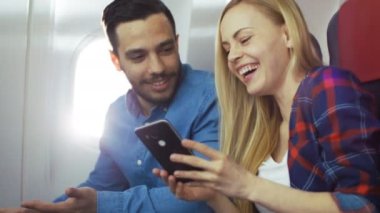 Ticari uçak Beautiful Genç bir gemide sarışın gösterir onun Smartphone perde yakışıklı İspanyol erkek için onlar hem gülmek. Uçak pencereden güneş parlıyor.