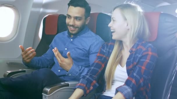 在一次商业飞机航班上英俊的西班牙裔男人给他美丽的金发女友讲有趣的故事。大家都笑了。他们在新的飞机，窗外阳光明媚旅行. — 图库视频影像