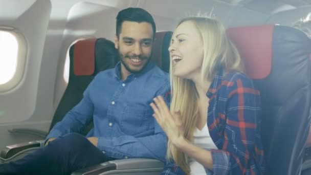 En un avión comercial El guapo hombre hispano cuenta una historia divertida a su hermosa novia rubia. Ambos se ríen. Viajan en avión nuevo, con el sol brillando a través de la ventana . — Vídeos de Stock