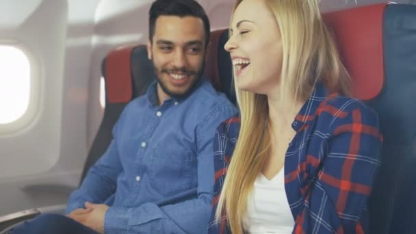 Na lot samolotem komercyjnych przystojny mężczyzna hiszpanin opowiada zabawna historia do jego piękna dziewczyna Blondynka. Zarówno śmiać. Podróżują w nowy samolot z słońce świeciło przez okno. — Wideo stockowe