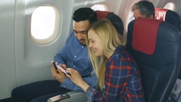 Na pokładzie samolotów komercyjnych piękna Młoda Blondynka z przystojny mężczyzna hiszpanin oglądać Social Media na smartfonie i śmiać się. Senior pasażera czyta książkę. — Wideo stockowe