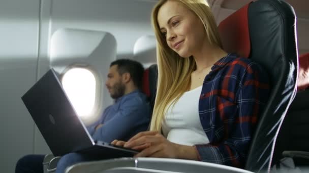 A bordo del avión comercial, la hermosa joven rubia trabaja en una computadora portátil mientras su vecino hispano masculino mira por la ventana . — Vídeo de stock
