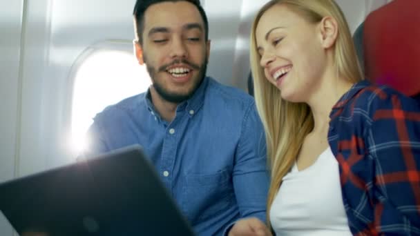 Na pokładzie samolotów komercyjnych piękna Młoda Blondynka z przystojny mężczyzna hiszpanin oglądać filmy na laptopie i śmiać się. Słońce świeci przez okno samolotu. — Wideo stockowe