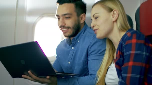 Op een bord van commerciële vliegtuig prachtige jonge Blonde met een knappe Spaanse mannelijke films op een Laptop en een lach. Zon schijnt door vliegtuig raam. — Stockvideo