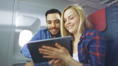 Ticari uçak bir gemide güzel genç sarışın yakışıklı İspanyol erkek kullanımı ile Tablet bilgisayar ve gülümseme. Uçak pencereden güneş parlıyor.
