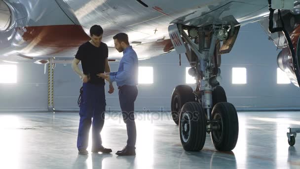 In einem Hangar zeigt ein Flugzeugwartungsingenieur dem Flugzeugtechniker technische Daten auf einem Tablet-Computer. Sie stehen in der Nähe eines sauberen nagelneuen Flugzeugs. — Stockvideo