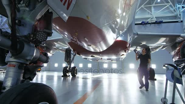 En un hangar Ingeniero / Técnico / Mecánico de Mantenimiento de Aeronaves inspecciona visualmente el chasis y el cuerpo / fuselaje del avión que camina debajo de él . — Vídeo de stock