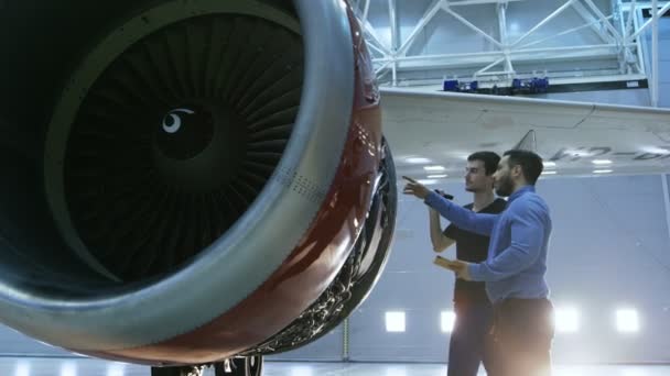 In einem Hangar zeigt ein Flugzeugwartungsingenieur dem Flugzeugtechniker technische Daten auf einem Tablet-Computer, er diagnostiziert das Triebwerk durch eine offene Luke. Sie stehen in der Nähe eines sauberen nagelneuen Flugzeugs. — Stockvideo