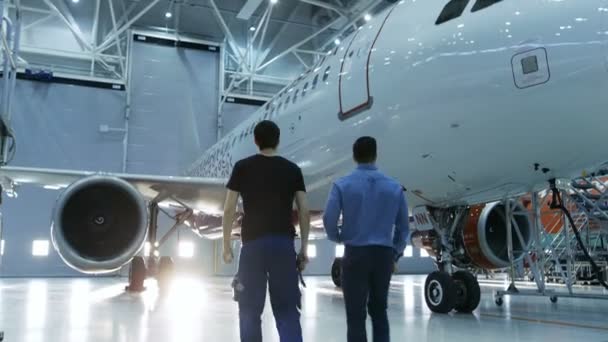 In einem Hangar zeigt ein Flugzeugwartungsingenieur dem Flugzeugtechniker technische Daten auf einem Tablet-Computer. Sie laufen neben sauberen nagelneuen Flugzeugen. — Stockvideo
