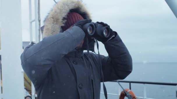 Aventurero en chaqueta caliente de pie en el barco y mirando a través de prismáticos. Es nevado y ventoso — Vídeos de Stock