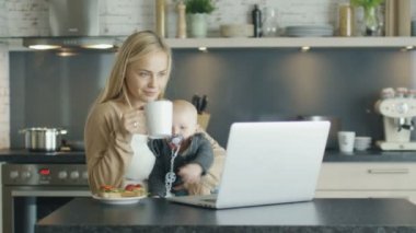 Laptop ve diz Enquisitive çocuk tutan önce mutfak masasının üzerinde gülümseyen anne oturur. Anne Sips--dan A Kupası çocuk uzanıyor Too.