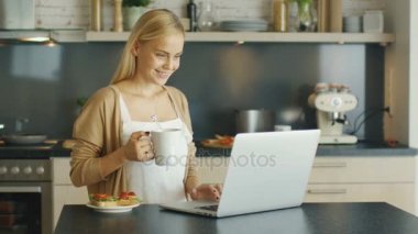 Güzel sarışın kadın mutfakta Laptop, o gülümsüyor ve bir kupa içecekler önce anlamına gelir. Ocakta yemek bir şey.