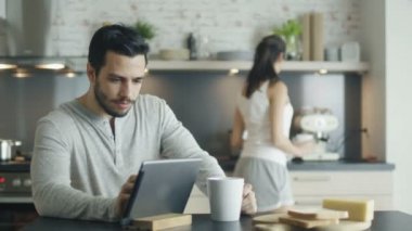 Mutfak genç adam içecek bir fincan kullanarak Tablet tamamlayın. Kız arkadaşı çıkageldi ve ona sarılarak.