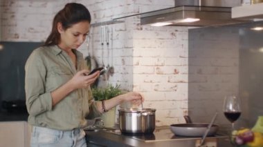 Genç kadın gıda kıpırdanırken tavada onun Smartphone tutarken ve gülümseyen.