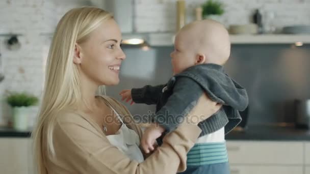 Joven madre sostiene y juega con su bebé mientras está de pie en la cocina — Vídeo de stock