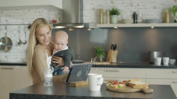 Ung mamma håller sin barn och använder surfplatta samtidigt på köket. — Stockvideo
