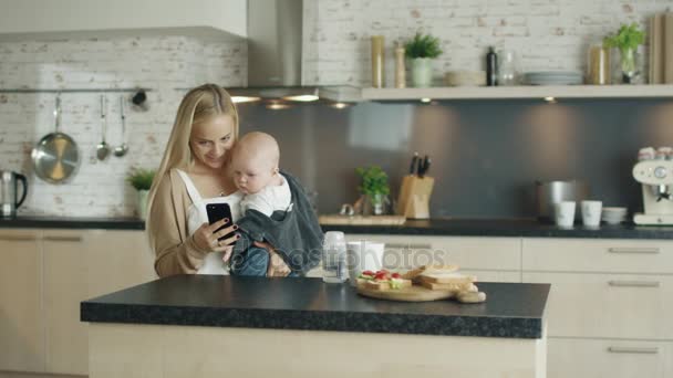 Giovane madre tiene il suo bellissimo bambino mentre è in cucina. Utilizza anche il suo smartphone con una mano, Bambino sembra interessato — Video Stock