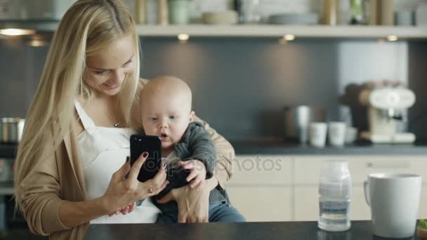 若い母親は、台所で彼女の赤ん坊を保持しています。彼女も使用してスマート フォンをやり取りするかわいい赤ちゃんに. — ストック動画