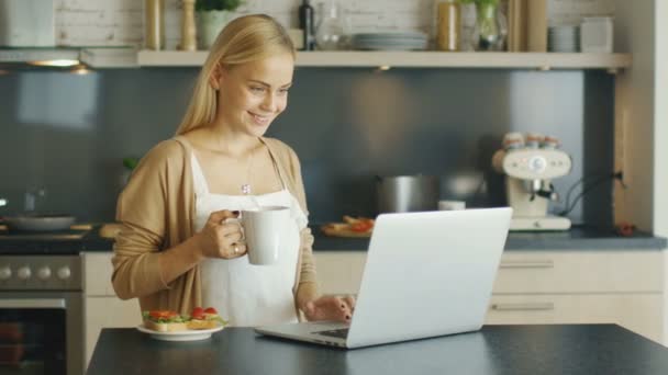 Härlig blond kvinna står i köket innan Laptop, hon ler och dricka från en mugg. Något är matlagning på spisen. — Stockvideo