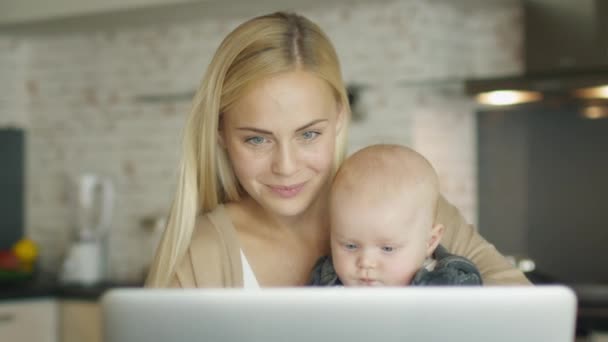 Сидя на кухне перед ноутбуком Улыбающаяся мать держит ребенка в руке и пьет из чашки . — стоковое видео